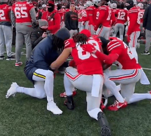 Players from opposing Ohio State and Michigan teams kneel together in solidarity on the field, demonstrating camaraderie post-game.