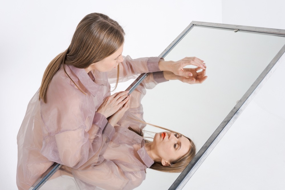 A woman in a pink shirt gazes at her reflection in a mirror, examining her appearance thoughtfully.