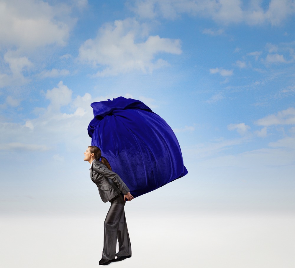 Businesswoman in a suit lifts a large purple bag against a clear sky, symbolizing burdens or challenges in a professional context.